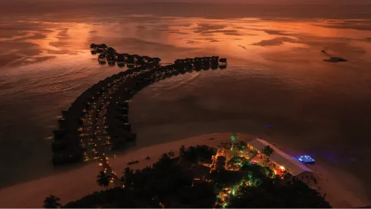 Curved overwater villa at Vakkaru Maldives illuminated with festive lights against a golden sunset ocean