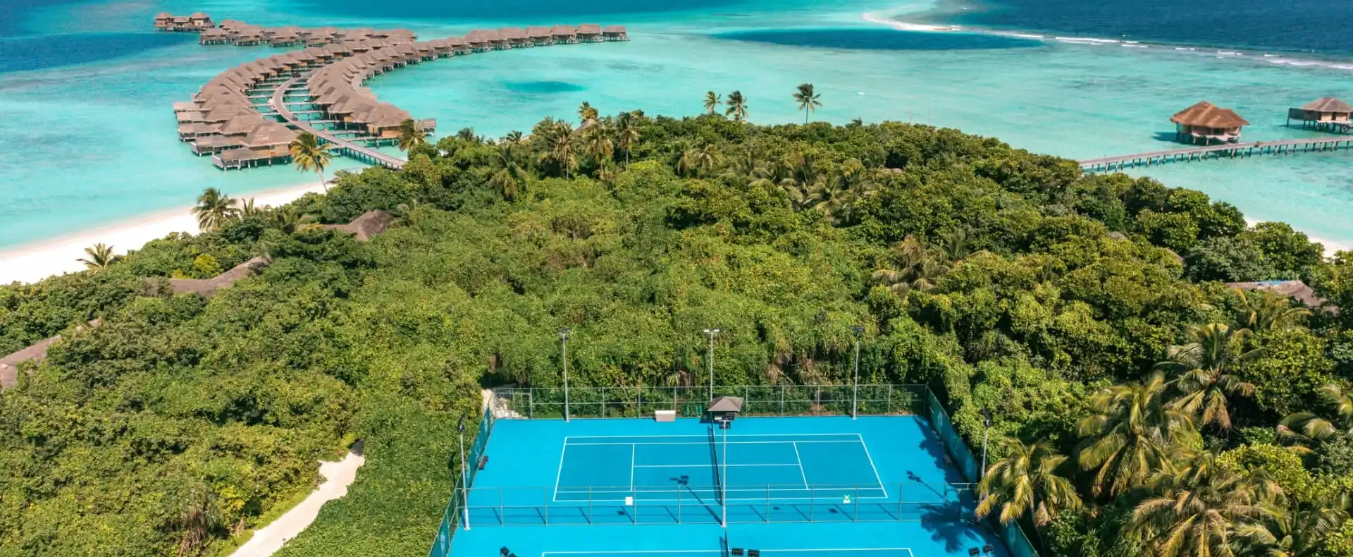 A vibrant blue tennis court is nestled within a lush green tropical island, flanked by white sand beaches and overwater bungalows curving across a turquoise ocean.