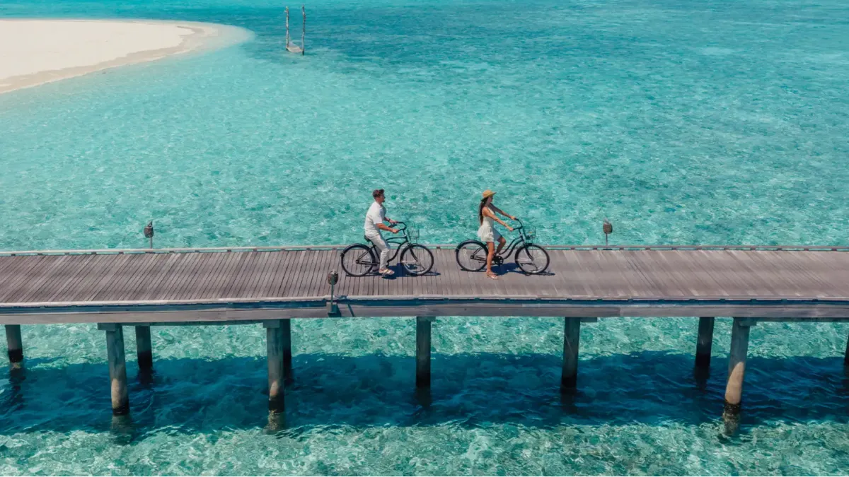 Couple cycling along a wooden jetty at Vakkaru Maldives above crystal-clear waters and island scenery.
