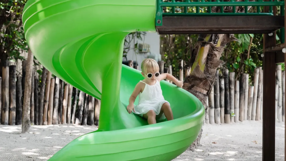 Child in white dress and sunglasses sliding down a green spiral slide in a tropical play area at Vakkaru Maldives.